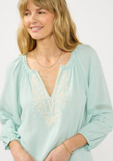 [Color: Dusty Mint] A front facing image of a blonde model wearing a mint green boho blouse with embroidered and lace details. 