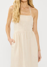 [Color: Natural] A detail image of a blonde model wearing a beige linen blend maxi dress with a halter neck, side pockets, and a back zip up closure. 