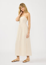 [Color: Natural] A side facing image of a blonde model wearing a beige linen blend maxi dress with a halter neck, side pockets, and a back zip up closure. 