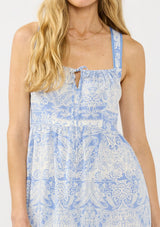 [Color: Periwinkle/White] A detail image of a blonde model wearing a white and blue boho maxi dress with adjustable straps, a square neckline with tie detail, a cross back, and side pockets. 