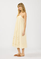 [Color: Cream] A side facing image of a blonde model wearing a flowy cutout embroidered midi dress with spaghetti straps and a back button detail. 