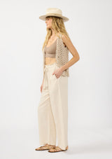 [Color: Natural] A side facing image of a blonde model wearing a beige crochet vest, tank top, and wide-leg pants with a drawstring waist and side pockets.