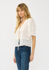[Color: White] A side facing image of a blonde model wearing a short sleeve top with embroidered eyelet, an open front with ties, and a v neckline.  