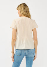 [Color: Natural] A back facing image of a blonde model wearing a lightweight beige top with embroidered cutouts, pintucks, and a v neckline.