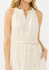 [Color: Natural] A detail image of a blonde model wearing a beige bohemian sleeveless maxi dress with a button front top, waist belt, side pockets, and contrast stitching. 