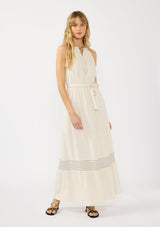 [Color: Natural] A front facing image of a blonde model wearing a beige bohemian sleeveless maxi dress with a button front top, waist belt, side pockets, and contrast stitching. 