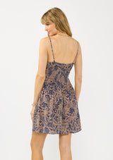 [Color: Navy/Natural] A back facing image of a blonde model wearing bohemian floral print mini dress with spaghetti straps, a smocked waist, a scoop neckline with tie detail, and side pockets. 