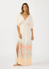 [Color: Natural/Coral] A front facing image of brunette model wearing a kimono sleeve maxi dress with a coral floral border print, smocked waist, a v-neckline, and side slits.