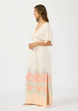 [Color: Natural/Coral] A side facing image of brunette model wearing a kimono sleeve maxi dress with a coral floral border print, smocked waist, a v-neckline, and side slits.