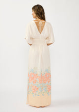 [Color: Natural/Coral] A back facing image of brunette model wearing a kimono sleeve maxi dress with a coral floral border print, smocked waist, a v-neckline, and side slits.