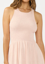 [Color: Shell Pink] A detail image of a brunette model wearing a sleeveless smocked midi dress with a round neck, side pockets, and a cross back with tie detail. 