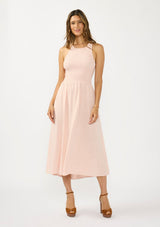 [Color: Shell Pink] A front facing image of a brunette model wearing a sleeveless smocked midi dress with a round neck, side pockets, and a cross back with tie detail. 
