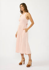 [Color: Shell Pink] A side facing image of a brunette model wearing a sleeveless smocked midi dress with a round neck, side pockets, and a cross back with tie detail. 