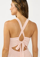 [Color: Shell Pink] A back facing image of a brunette model wearing a sleeveless smocked midi dress with a round neck, side pockets, and a cross back with tie detail. 