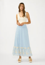 [Color: Chambray Blue/Natural] A front facing image of a brunette model wearing a light blue maxi skirt with a smocked waist and embroidered hem. 