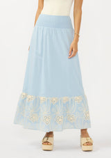 [Color: Chambray Blue/Natural] A front facing image of a light blue maxi skirt with a smocked waist and embroidered hem. 