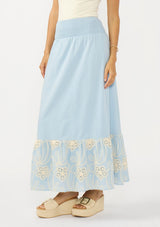 [Color: Chambray Blue/Natural] A side facing image of a light blue maxi skirt with a smocked waist and embroidered hem. 