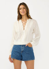 [Color: Natural] A front facing image of a brunette model wearing a white textured cotton blouse with a lace up front, lace insert, and long sleeves with dropped shoulders. 
