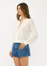[Color: Natural] A side facing image of a brunette model wearing a white textured cotton blouse with a lace up front, lace insert, and long sleeves with dropped shoulders. 