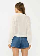 [Color: Natural] A back facing image of a brunette model wearing a white textured cotton blouse with a lace up front, lace insert, and long sleeves with dropped shoulders. 