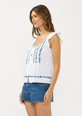 [Color: White/Blue] A side facing image of a brunette model wearing a white flutter sleeve top with blue floral embroidery, a button front, and pintucks. 