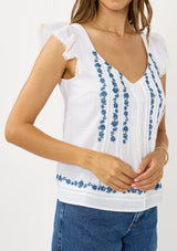 [Color: White/Blue] A detail image of a brunette model wearing a white flutter sleeve top with blue floral embroidery, a button front, and pintucks. 