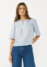 [Color: Blue] A front facing image of a brunette model wearing a light blue cotton blouse with a ruffled yoke, half sleeves, and a button front.