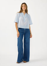 [Color: Blue] A front facing image of a brunette model wearing a light blue cotton blouse with a ruffled yoke, half sleeves, and a button front.