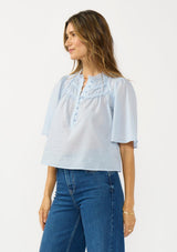 [Color: Blue] A side facing image of a brunette model wearing a light blue cotton blouse with a ruffled yoke, half sleeves, and a button front.