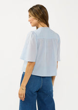 [Color: Blue] A back facing image of a brunette model wearing a light blue cotton blouse with a ruffled yoke, half sleeves, and a button front.