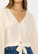 [Color: Tea Dye] A detail image of a brunette model wearing a light beige blouse with wide long sleeves, a v neckline, button front, and a tie front detail. 
