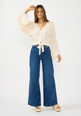 [Color: Tea Dye] A front facing image of a brunette model wearing a light beige blouse with wide long sleeves, a v neckline, button front, and a tie front detail. 