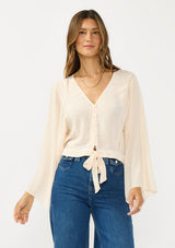 [Color: Tea Dye] A front facing image of a brunette model wearing a light beige blouse with wide long sleeves, a v neckline, button front, and a tie front detail. 