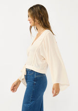 [Color: Tea Dye] A side facing image of a brunette model wearing a light beige blouse with wide long sleeves, a v neckline, button front, and a tie front detail. 