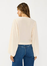 [Color: Tea Dye] A back facing image of a brunette model wearing a light beige blouse with wide long sleeves, a v neckline, button front, and a tie front detail. 
