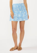 [Color: Blue/Ivory] A front facing image of a brunette model wearing a white bodysuit with a blue floral smocked mini skirt with tiered layers. 
