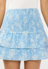 [Color: Blue/Ivory] A detail image of a brunette model wearing a white bodysuit with a blue floral smocked mini skirt with tiered layers. 