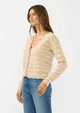 [Color: Heather Sand/White] A side facing image of a brunette model wearing a light brown pointelle sweater with a v-neckline, button front, and a scalloped trim. 