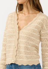 [Color: Heather Sand/White] A detail image of a brunette model wearing a light brown pointelle sweater with a v-neckline, button front, and a scalloped trim. 