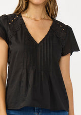 [Color: Black] A detail image of a brunette model wearing a lightweight black top with embroidered cutouts, pintucks, and a v neckline. 