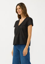 [Color: Black] A side facing image of a brunette model wearing a lightweight black top with embroidered cutouts, pintucks, and a v neckline. 