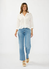 [Color: White] A front facing image of a brunette model wearing a white striped blouse with a button front, collared neck, and a high low hemline with ruffle trim.