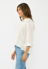 [Color: White] A side facing image of a brunette model wearing a white striped blouse with a button front, collared neck, and a high low hemline with ruffle trim.