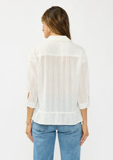 [Color: White] A back facing image of a brunette model wearing a white striped blouse with a button front, collared neck, and a high low hemline with ruffle trim.
