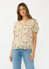 [Color: Blush/Rose] A front facing image of a brunette model wearing a pink and green floral v neck top with a button front and a ruffle trim accent. 
