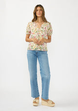 [Color: Blush/Rose] A front facing image of a brunette model wearing a pink and green floral v neck top with a button front and a ruffle trim accent. 