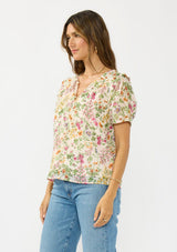 [Color: Blush/Rose] A side facing image of a brunette model wearing a pink and green floral v neck top with a button front and a ruffle trim accent. 