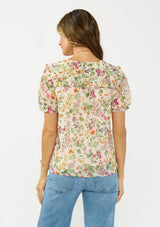 [Color: Blush/Rose] A back facing image of a brunette model wearing a pink and green floral v neck top with a button front and a ruffle trim accent. 
