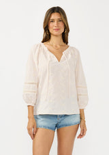 [Color: Tan/Natural] A front facing image of a brunette model wearing a beige checkered blouse with floral embroidery, lace details, and a split v neckline with ties. 