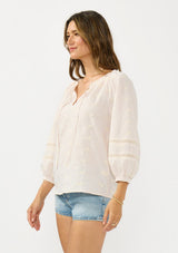 [Color: Tan/Natural] A side facing image of a brunette model wearing a beige checkered blouse with floral embroidery, lace details, and a split v neckline with ties. 